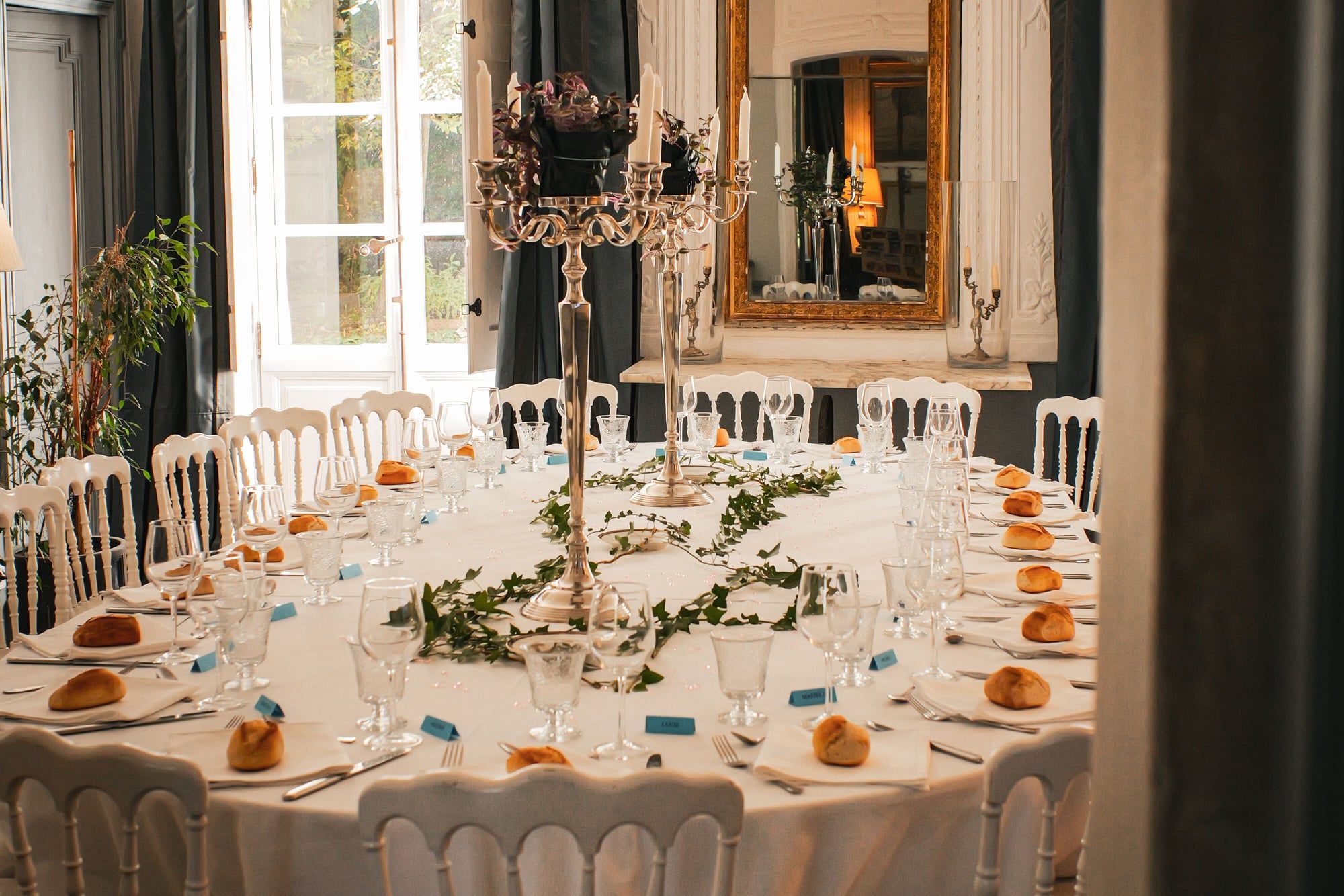 Grande table ronde dressée dans un salon à moulures, chandeliers et chaises blanches